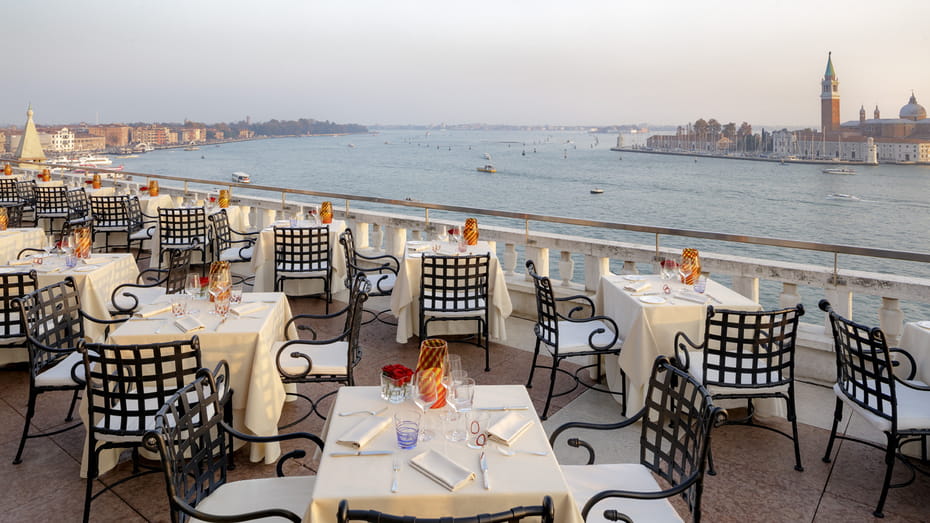 The rooftop terrace of Restaurant Terrazza Danieli at sunset. Small, square, candlelit tables dressed in tablecloths epitomise fine dining in Venice. Spectacular views overlook the Venetian lagoon with tradtional gondolas.