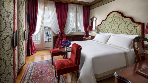 A luxury lagoon view room at Hotel Danieli in Venice. The restyled design by Jacques Garcia illustrates the rich drama of the Gothic era. Beautiful Venetian lagoon views can be enjoyed from a private balcony accessed from glass doors. There is an elegant king sized bed with white linen and a green and gold headboard. There are red velvet chairs, herringbone flooring and large wardrobes for storage.