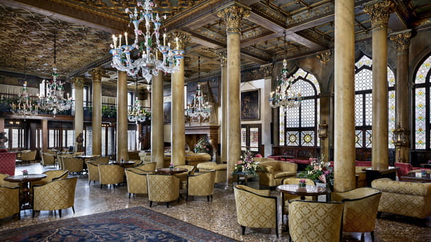 A luxury lagoon view room at Hotel Danieli in Venice. The restyled design by Jacques Garcia illustrates the rich drama of the Gothic era. Beautiful Venetian lagoon views can be enjoyed from a private balcony accessed from glass doors. There is an elegant king sized bed with white linen and a green and gold headboard. There are red velvet chairs, herringbone flooring and large wardrobes for storage.